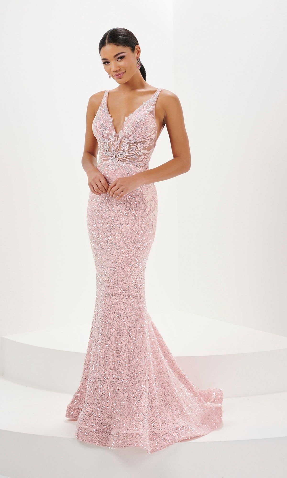 A woman stands on a white platform wearing the Tiffany Sequin Velvet Mermaid Prom Dress 16110, a fitted, sleeveless light pink gown with sequin floral appliques, deep V neckline, backless design, and a subtle train.